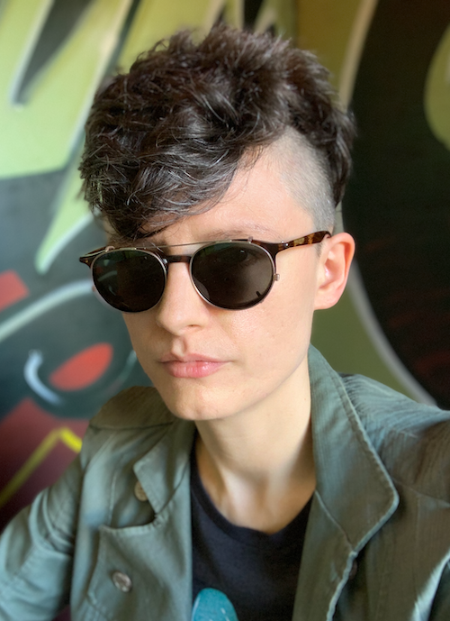 A white person with short curly brown hair and glasses wearing sunglasses and a green jacket.