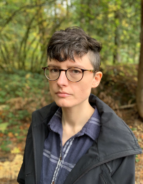 A white person with short curly brown hair and glasses wearing a blue flannel shirt and black rain jacket.